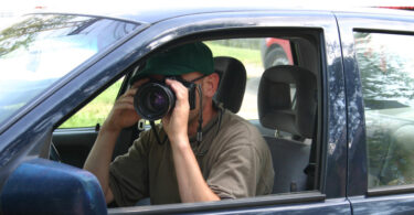 Detektiv in einem Auto, der aus dem Seitenfenster heraus ein Foto schießt.