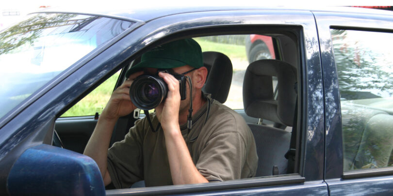 Detektiv in einem Auto, der aus dem Seitenfenster heraus ein Foto schießt.