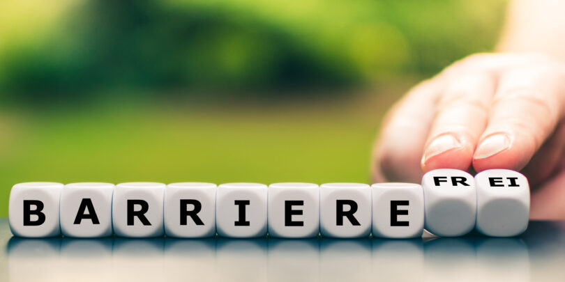Nahaufnahme einer Hand, die in einer Reihe beschrifteter Würfeln zwei davon umdreht und so das Wort "Barriere" in "barrierefrei" ändert.