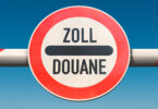 Schranke, in deren Mitte ein rundes, rot umrandetes Schild mit der Aufschrift Zoll-Douane (schwarze Schrift auf weißem Grund) angebracht ist.