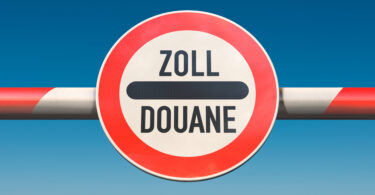 Schranke, in deren Mitte ein rundes, rot umrandetes Schild mit der Aufschrift Zoll-Douane (schwarze Schrift auf weißem Grund) angebracht ist.
