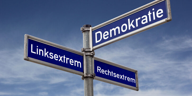 Verkehrsschild für Straßen: Es hat drei Straßennamen. Auf einem steht Demokratie, auf dem anderen Linksextrem und auf dem letzten Rechtsextrem.