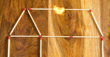 Haus aus Zündhölzern, das auf eine Tischplatte gelegt wurde und bei dem der Dachbalken in Flammen steht.