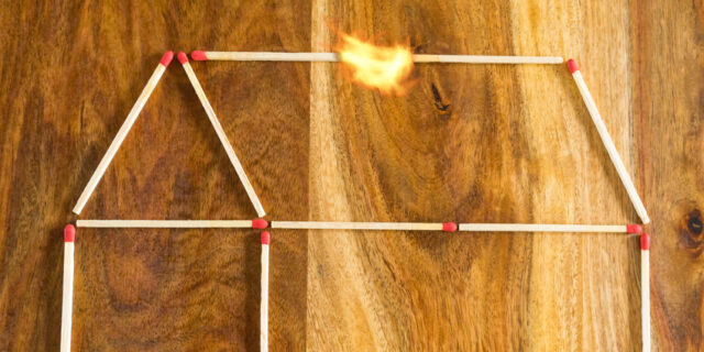 Haus aus Zündhölzern, das auf eine Tischplatte gelegt wurde und bei dem der Dachbalken in Flammen steht.