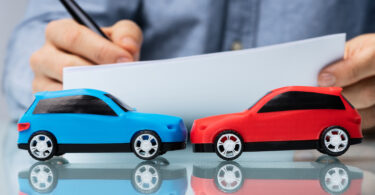 Nahaufnahme von zwei Spielzeugautos, die frontal zusammenstoßen. Im Hintergrund sind die Hände eines Mannes zu sehen, der etwas auf ein Blatt Papier schreibt.