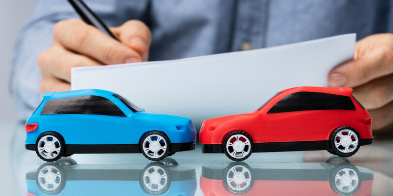 Nahaufnahme von zwei Spielzeugautos, die frontal zusammenstoßen. Im Hintergrund sind die Hände eines Mannes zu sehen, der etwas auf ein Blatt Papier schreibt.