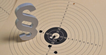 Bildmontage, in der auf einer papiernen Zielscheibe mit Einschusslöchern ein graues Paragrafen-Zeichen steht.