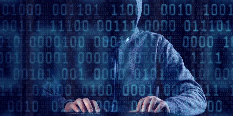 Person mit Kapuze, deren Gesicht dadurch nicht zu erkennen ist, die an einer Tastatur sitzt. Im Vordergrund des Bildes befindet sich eine blaue Zahlenmatrix. Der Hintergrund ist schwarz gehalten.