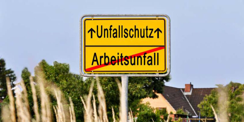 Gelbe Ortstafel, in deren unterer Hälfte ein rot durchgestrichenes 'Arbeitsunfall' steht. In der oberen Hälfte steht hingegen das Wort 'Unfallschutz'. Links und rechts von diesem befindet sich jeweils ein Pfeil, der nach oben zeigt.