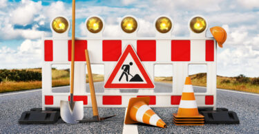 Baustelle, Absperrung, Straßensperrung mit Schaufel, Pickel, Baustellenschild, Pylone und Sicherheitshelm auf Straße.