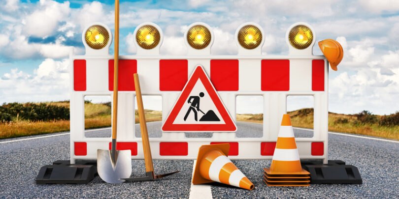 Baustelle, Absperrung, Straßensperrung mit Schaufel, Pickel, Baustellenschild, Pylone und Sicherheitshelm auf Straße.