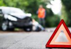 A close up of a red emergency triangle on the road in front of a damaged car and unrecognizable people.