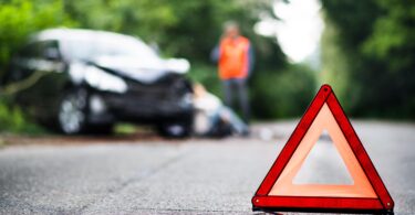 A close up of a red emergency triangle on the road in front of a damaged car and unrecognizable people.