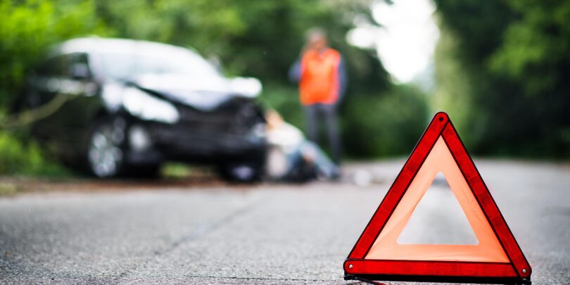 A close up of a red emergency triangle on the road in front of a damaged car and unrecognizable people.