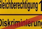 Gelbes Schild, dass durch einen horizontalen schwarzen Strich in zwei Hälften geteilt wird. In der unteren Hälfte steht das Wort "Diskriminierung". Es ist mit einem diagonalen roten Strich durchgestrichen. In der oberen Hälfte steht das Wort "Gleichberechtigung". Rechts daneben befindet sich ein nach oben zeigender schwarzer Pfeil.