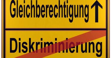 Gelbes Schild, dass durch einen horizontalen schwarzen Strich in zwei Hälften geteilt wird. In der unteren Hälfte steht das Wort "Diskriminierung". Es ist mit einem diagonalen roten Strich durchgestrichen. In der oberen Hälfte steht das Wort "Gleichberechtigung". Rechts daneben befindet sich ein nach oben zeigender schwarzer Pfeil.