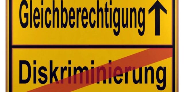 Gelbes Schild, dass durch einen horizontalen schwarzen Strich in zwei Hälften geteilt wird. In der unteren Hälfte steht das Wort "Diskriminierung". Es ist mit einem diagonalen roten Strich durchgestrichen. In der oberen Hälfte steht das Wort "Gleichberechtigung". Rechts daneben befindet sich ein nach oben zeigender schwarzer Pfeil.
