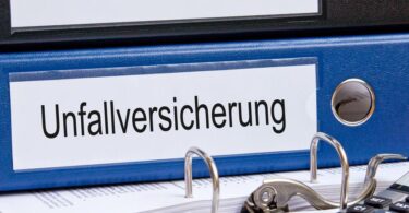 Blauer Aktenordner, der auf einem Schreibtisch liegt und auf dessen Rückenschild "Unfallversicherung" steht.