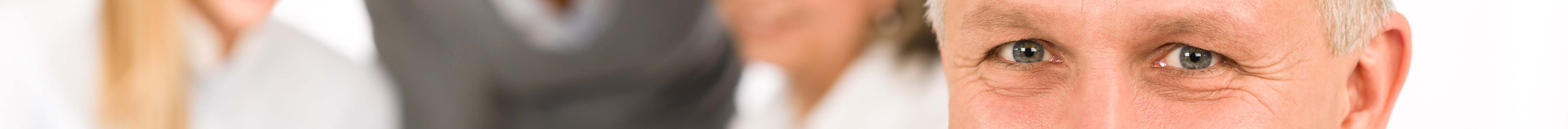 Ein älterer Mann in einem hellblauen Hemd und einer blauen Krawatte lächelt in die Kamera, während er vor einem Laptop steht. Im Hintergrund sind drei unscharfe Personen zu sehen, die an einem Laptop arbeiten.