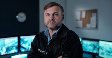 Sicherheitsmitarbeiter in schwarzer Jacke mit Aufschrift 'SECURITY' hält Funkgerät in einem Kontrollraum mit mehreren Monitoren.