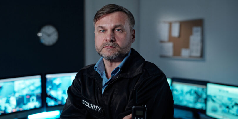 Sicherheitsmitarbeiter in schwarzer Jacke mit Aufschrift 'SECURITY' hält Funkgerät in einem Kontrollraum mit mehreren Monitoren.