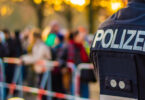 Eine Aufnahme von hinten zeigt einen Polizisten in Uniform mit der Aufschrift „POLIZEI“ auf dem Rücken, während im Hintergrund eine verschwommene Menschenmenge hinter Absperrbändern steht, vermutlich bei einer öffentlichen Veranstaltung.