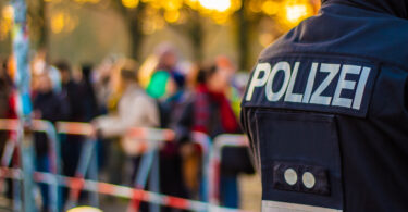 Eine Aufnahme von hinten zeigt einen Polizisten in Uniform mit der Aufschrift „POLIZEI“ auf dem Rücken, während im Hintergrund eine verschwommene Menschenmenge hinter Absperrbändern steht, vermutlich bei einer öffentlichen Veranstaltung.