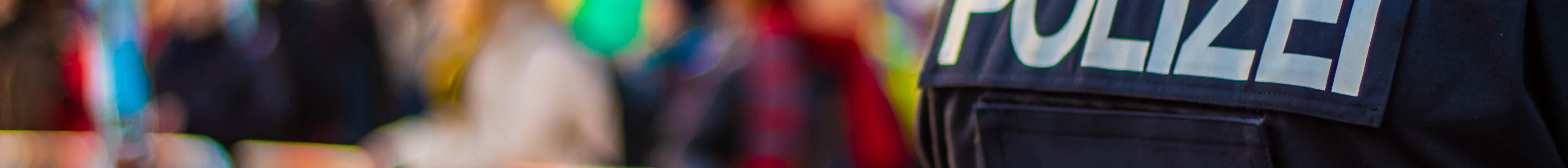 Eine Aufnahme von hinten zeigt einen Polizisten in Uniform mit der Aufschrift „POLIZEI“ auf dem Rücken, während im Hintergrund eine verschwommene Menschenmenge hinter Absperrbändern steht, vermutlich bei einer öffentlichen Veranstaltung.