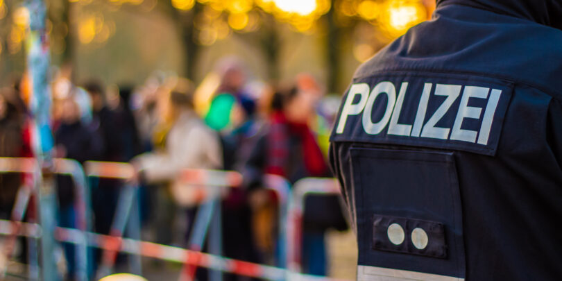Eine Aufnahme von hinten zeigt einen Polizisten in Uniform mit der Aufschrift „POLIZEI“ auf dem Rücken, während im Hintergrund eine verschwommene Menschenmenge hinter Absperrbändern steht, vermutlich bei einer öffentlichen Veranstaltung.