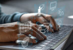 Nahaufnahme zweier Hände, die die Tastatur eines geöffneten Laptops bedienen. Über den Händen schweben folgende virtuelle Symbole: ein Kopf mit Zahnrädern darin, ein aufgeschlagenes Buch, eine Person, die ein Smartbord bedient, ein Doktorhut, ein Bildschirm, ein Headset, eine leuchtende Glühbirne.