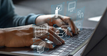 Nahaufnahme zweier Hände, die die Tastatur eines geöffneten Laptops bedienen. Über den Händen schweben folgende virtuelle Symbole: ein Kopf mit Zahnrädern darin, ein aufgeschlagenes Buch, eine Person, die ein Smartbord bedient, ein Doktorhut, ein Bildschirm, ein Headset, eine leuchtende Glühbirne.