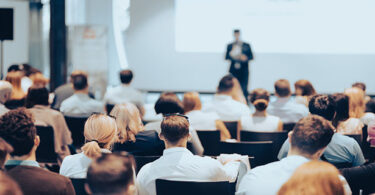 Referent, der einen Vortrag bei einer Geschäftsveranstaltung in einem Konferenzsaal hält. Rückansicht von nicht erkennbaren Personen im Publikum.