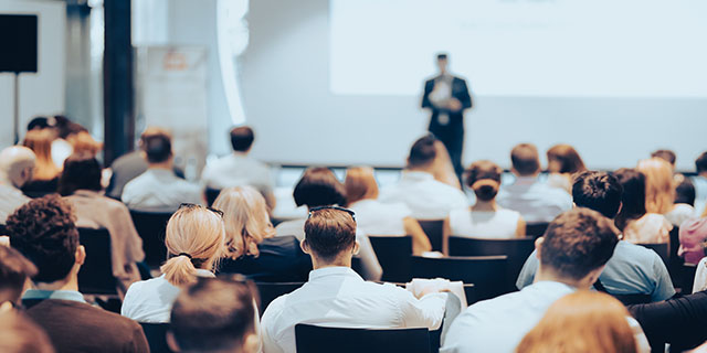 Referent, der einen Vortrag bei einer Geschäftsveranstaltung in einem Konferenzsaal hält. Rückansicht von nicht erkennbaren Personen im Publikum.
